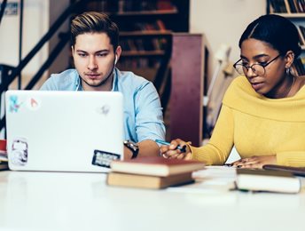 two students sitting at a desk with books and laptop