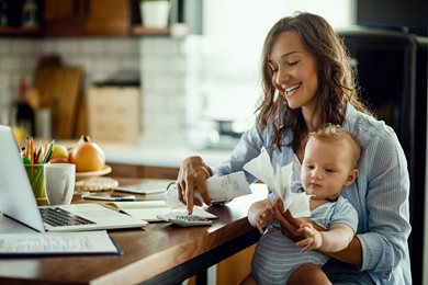 mother sitting at desk with baby, receipts, laptop and calculator