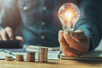 person on calculator holding lightbulb with coins staked on desk